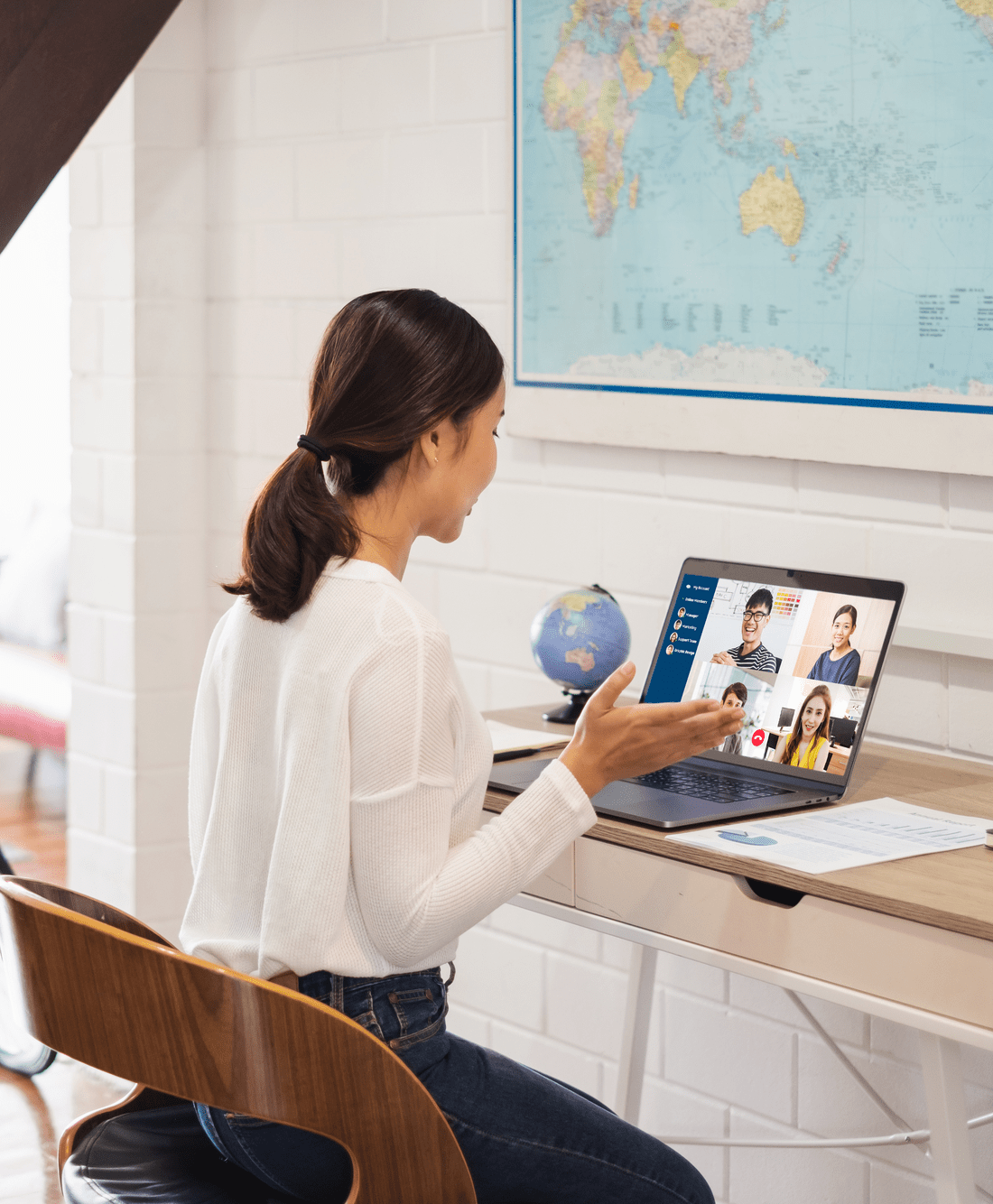 A woman in a white sweater participates in a virtual meeting on her laptop at a desk.